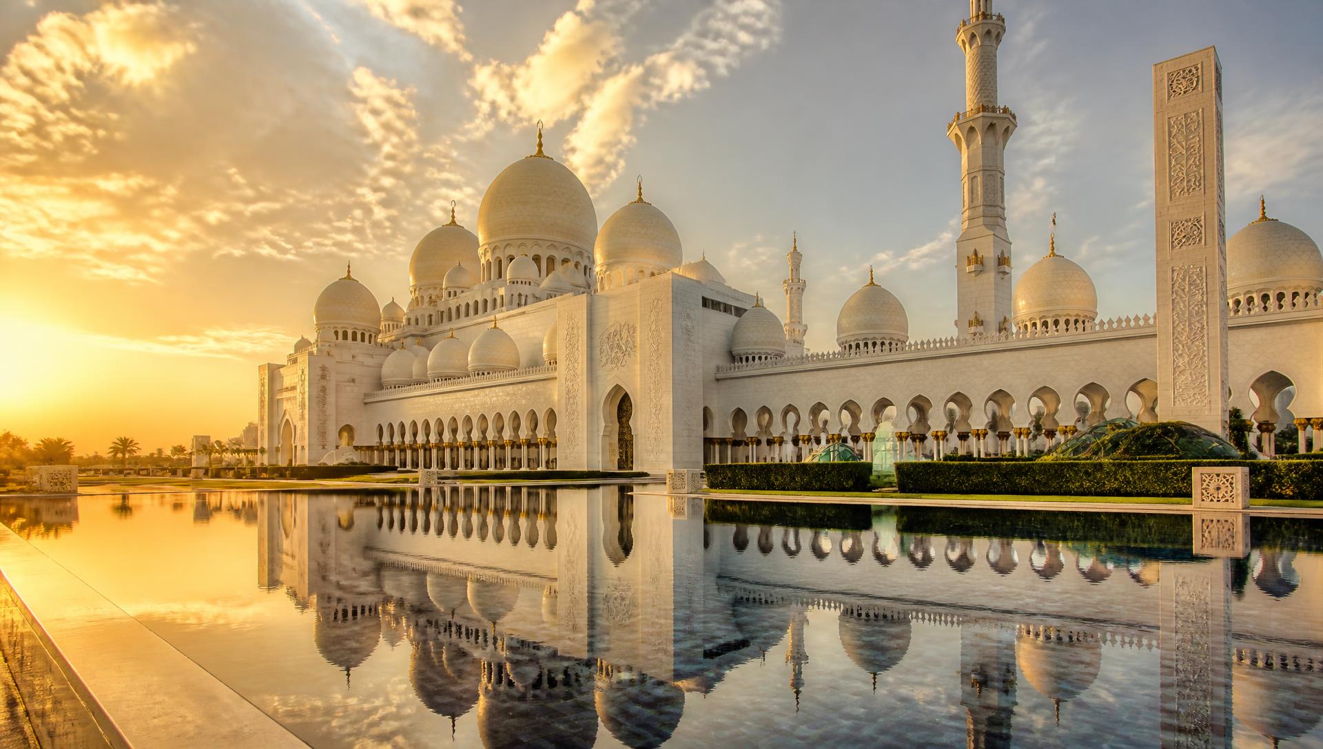 The white marble domes and minarets of Sheikh Zayed Grand Mosque in Abu Dhabi reflected in a courtyard pool at golden hour