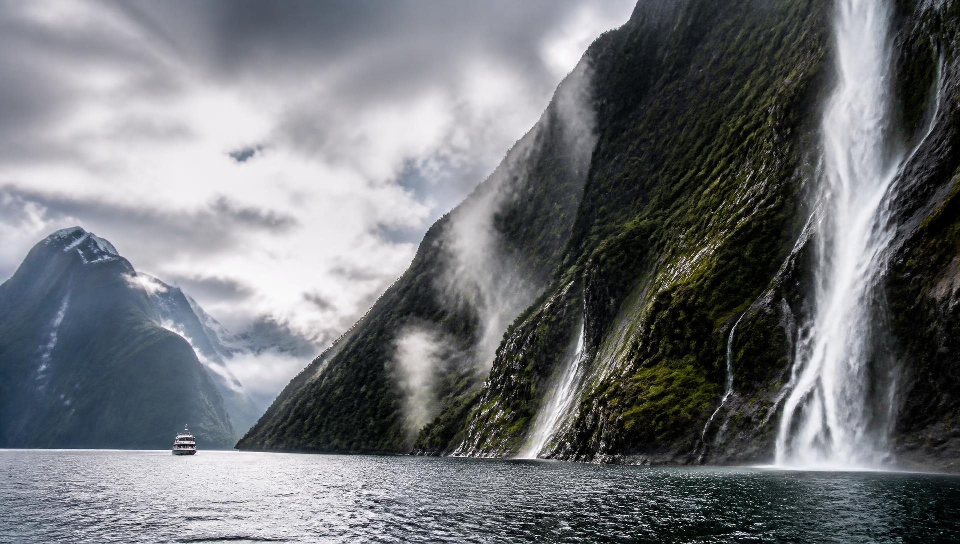 Waterfalls tumbling down steep cliffs into the dark water of Milford Sound under dramatic clouds